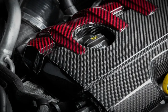 An engine cover made of carbon fiber, with red and black weave patterns, is shown within an engine compartment. A cap with yellow text is visible in the center.