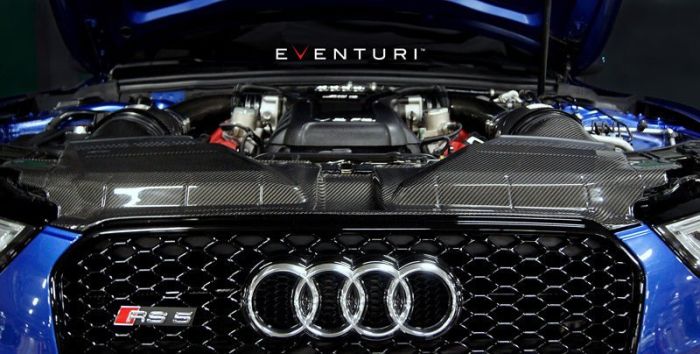 The blue Audi RS 5 showcases its exposed engine with "Eventuri" branding above, surrounded by carbon fiber components and the iconic Audi grille in a dimly lit setting.