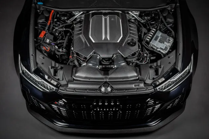 An intricately designed car engine with a carbon fiber cover sits inside the engine bay of a modern luxury car, surrounded by various cables and components, with the front grille partially visible.