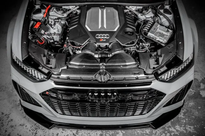 An Audi RS car's engine bay is displayed with a V8 engine, surrounded by connected hoses and components resting in a clean, gray car body in a garage.