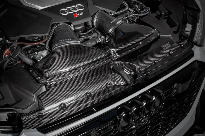 An Audi R8 engine with “Eventuri” intake components in a carbon fiber finish, situated in the engine bay of the car, showcasing the manufacturer's logo prominently.