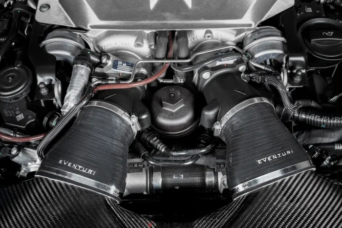 Engine components with "Eventuri" branded air intakes positioned centrally, surrounded by various cables and metal parts inside the car engine bay. The air intakes are covered by patterned carbon fiber material.