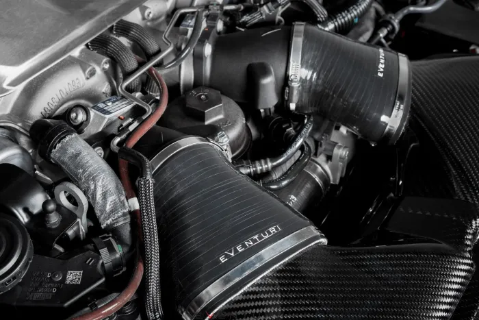 Eventuri-branded engine components, including black intake ducts and hoses, are fitted within a car engine bay, featuring metal parts, cables, and tubes intricately connected.