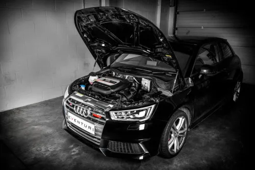 A black car with its hood open, showcasing the engine, is stationary in a dimly lit garage next to a white brick wall. Text on the license plate reads "EVENTURI".