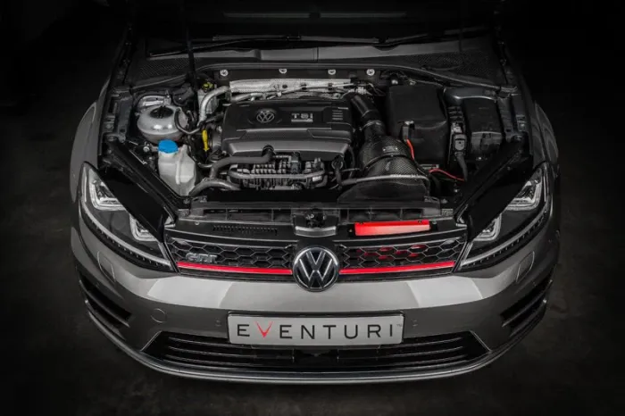 The engine compartment of a silver Volkswagen GTI is shown with the hood open, featuring a TSI engine. A white license plate reads "EVENTURI," and the car is in a dimly lit garage.