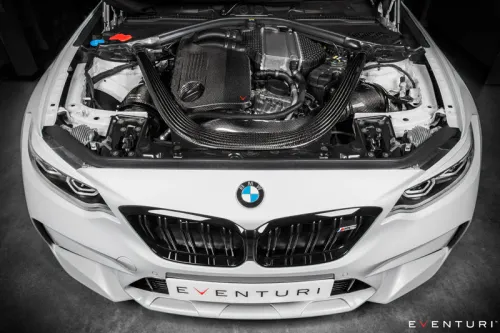 A BMW car's engine bay with a carbon fiber intake system, displayed in a garage with the hood open. The front grille features the BMW logo. The front plate reads "EVENTURI."