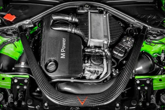 A carbon fiber engine cover labeled "M Power" sits between black hoses and metal components, surrounded by neon green car chassis and black wiring inside a vehicle's engine bay.