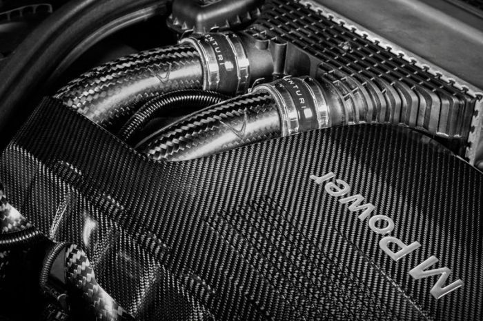Carbon fiber tubes connected to an engine covered by a carbon fiber panel inscribed with "M Power" in an automotive engine bay.