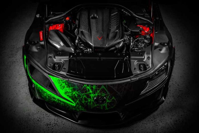 A sports car's hood is open, revealing a carbon fiber engine. The front bumper features vibrant green, jagged patterning. The car is stationary in a dimly lit garage.