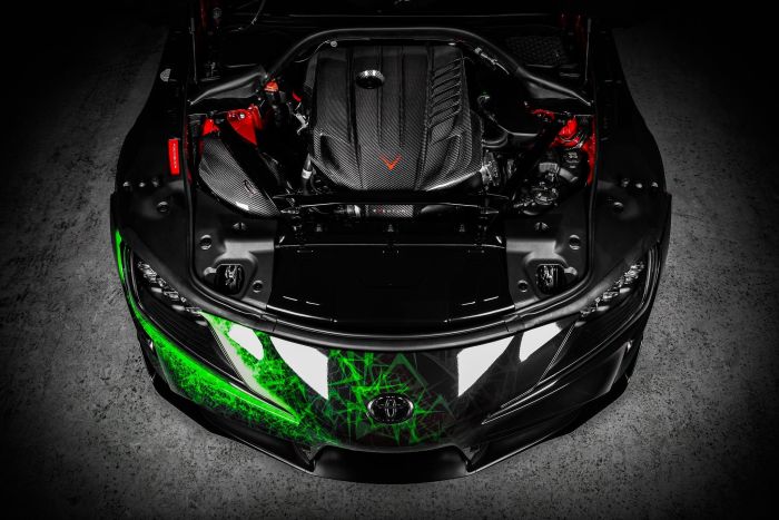 A car's engine bay with a detailed, carbon-fiber engine cover is displayed on a sleek black car, highlighted with green neon graphical patterns, situated on a concrete floor.