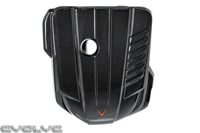 Black carbon fiber engine cover with a central circular opening and an orange V-shaped emblem, featuring ridged textures. "EVOLVE" text appears in the bottom-left corner.