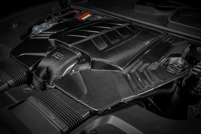 A carbon fiber engine cover with intricate patterns is fitted over the engine components and hoses inside the engine bay of a car.