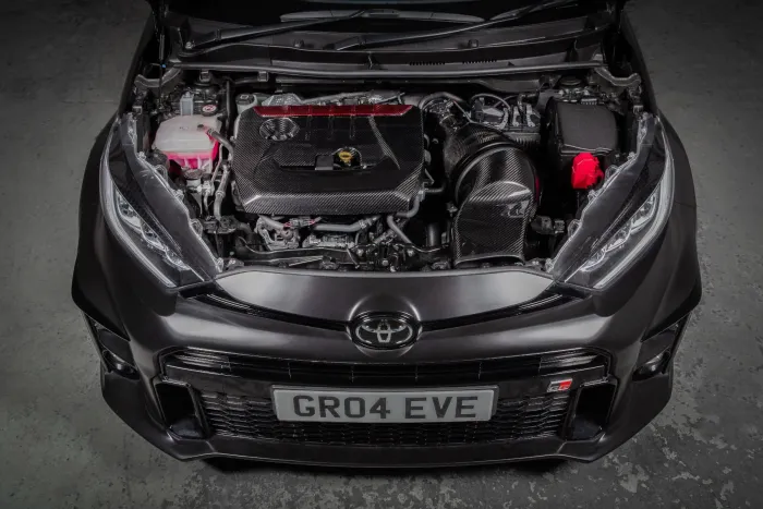 Car engine exposed under an open hood in a garage setting. The vehicle has a sleek design with visible Toyota emblem and license plate reads “GR04 EVE.”