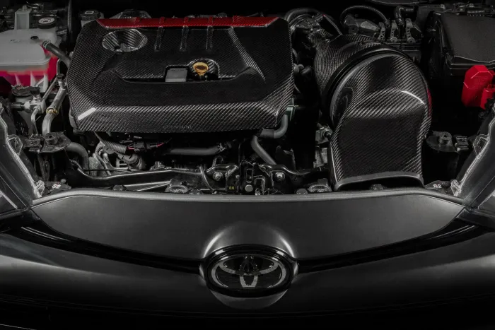 A car engine with a carbon fiber cover and components; the Toyota logo is visible in the foreground, surrounded by other mechanical parts and wires in the engine bay.