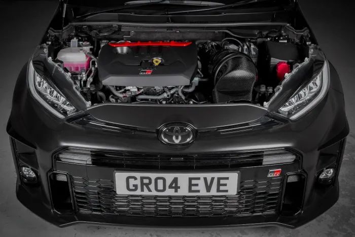Car with open hood displaying engine components, situated in a dimly lit garage. The front grille has a license plate reading "GR04 EVE" and a Toyota emblem.