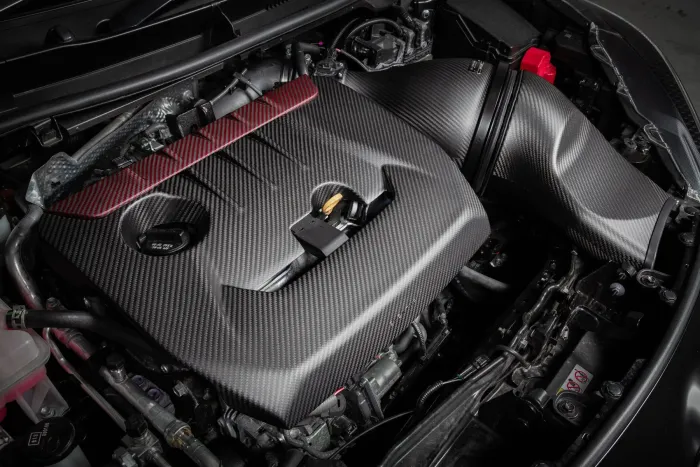 A carbon fiber engine cover sits atop a car engine, featuring sleek design and precise detailing in a workshop setting. Nearby components and wiring are visible, highlighting a well-maintained automotive system.