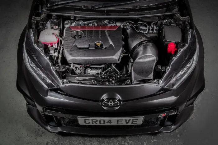 The opened hood reveals a well-kept car engine, with visible carbon fiber components, and "GR04 EVE" on the license plate, set in a workshop with a concrete floor.