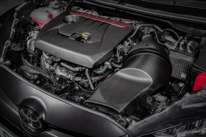 Car engine covered with a sleek, black carbon fiber cover; surrounded by various hoses, wires, and coolant tank, within a Toyota vehicle's engine compartment.