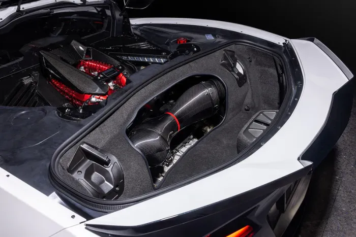 A high-performance car engine with red and black components is displayed in the engine bay of a white vehicle, surrounded by black and grey materials under bright lighting.