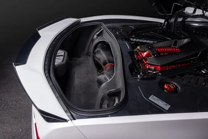 A white sports car, with its rear trunk open, exposing a red and black engine, and a small storage compartment lined with a black textured cover. The setting is dimly lit.