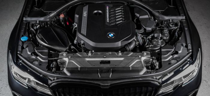 A BMW engine labeled "BMW M Power Turbo" sits within a sleek black car's open hood, surrounded by various components and mechanical parts.