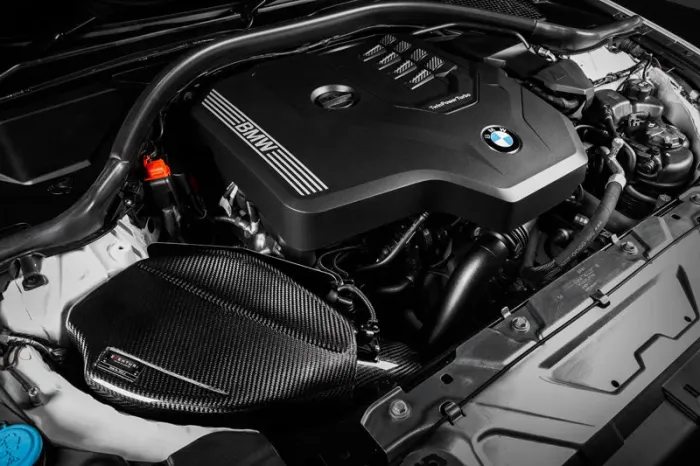 An engine with a black cover marked "BMW" and "TwinPower Turbo" is housed in a car's engine bay, surrounded by various mechanical components and tubing.