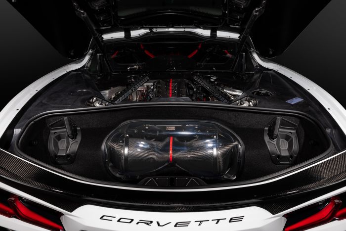 A Corvette with its rear engine compartment open, showcasing a carbon fiber engine component with a red stripe. The surrounding area features sleek, black detailing in a brightly lit environment. "CORVETTE" text is visible at the bottom.