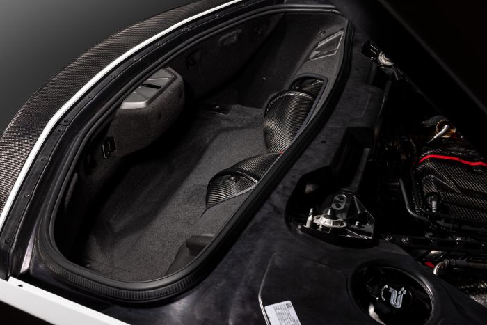Car trunk with black carbon fiber and gray interior, showing components and storage space. Surrounding the context is the interior of the car, partly showing an engine.