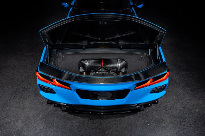 A blue sports car with its rear trunk open, revealing a carbon fiber component, is parked on a dark surface in a low-lit environment.