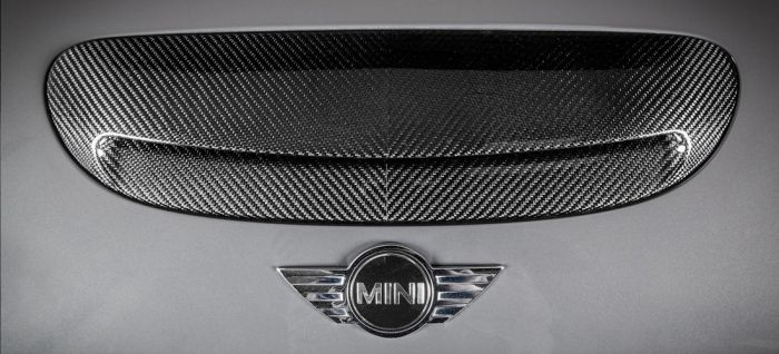 A carbon fiber air vent is mounted on a metallic surface, with the "MINI" emblem featuring wings situated below it. The metallic surface appears to be part of a vehicle.
