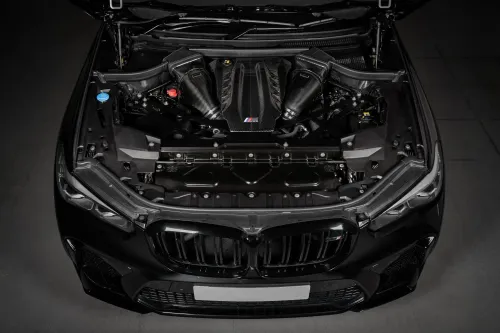 Car engine compartment (object) displayed in an open hood (action) of a black BMW vehicle, featuring carbon fiber components and the "M" logo on the central cover (context).