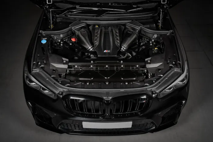 Car engine compartment (object) displayed in an open hood (action) of a black BMW vehicle, featuring carbon fiber components and the "M" logo on the central cover (context).