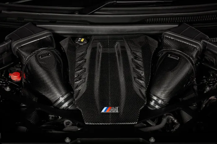 A carbon fiber engine cover, displaying a red, blue, and light blue-striped "M" logo, is situated within a car's polished engine bay.