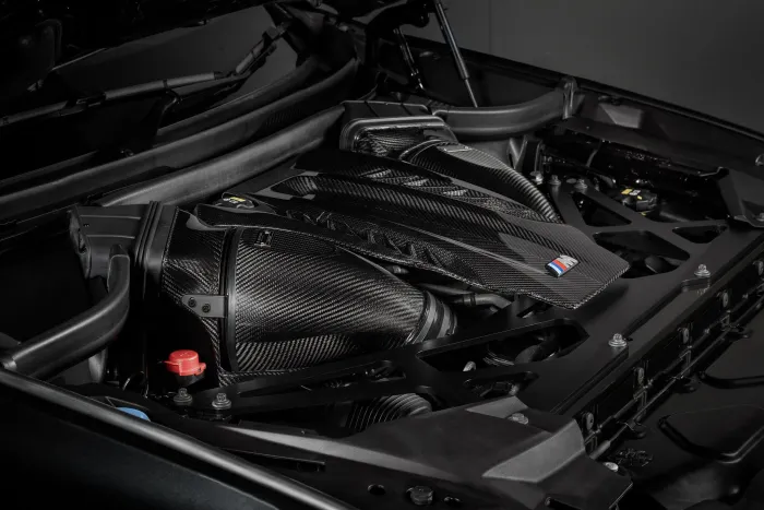 Carbon fiber car engine cover on a high-performance engine, featuring a blue, violet, and red "M" logo, set within a dark mechanical environment.