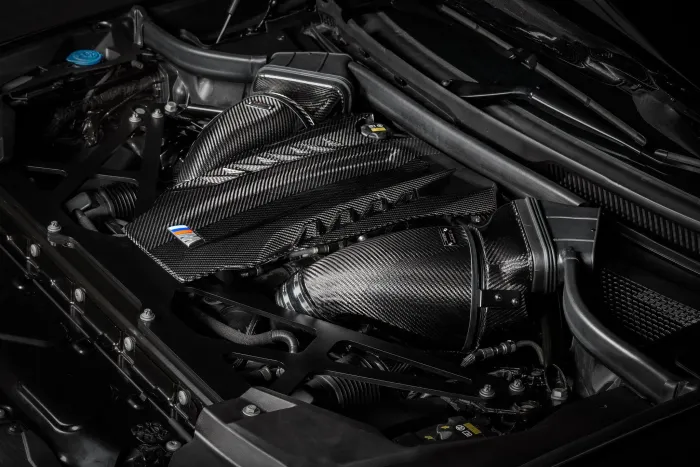 Carbon fiber engine cover and air intakes, all adorned with a small colored emblem, situated in the sleek engine bay of a modern car.