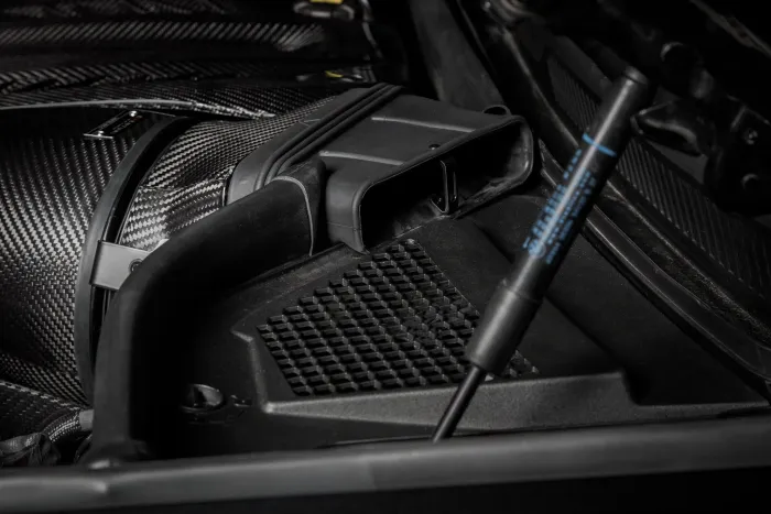 Carbon fiber intake system connected to the air filter housing, situated within the hood compartment of a vehicle. The compartment features various black components and a hydraulic rod labeled "Stabilus."