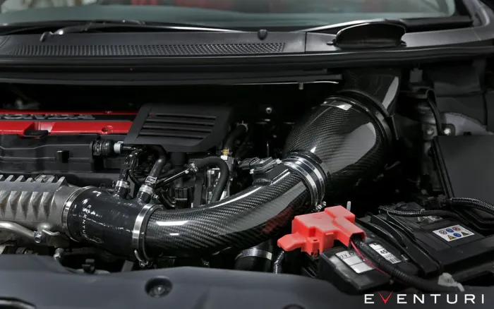 Carbon fiber air intake system installed in a car engine bay, with red components and a visible battery, surroundings include tubes and other engine parts. Text reads: "EVENTURI."