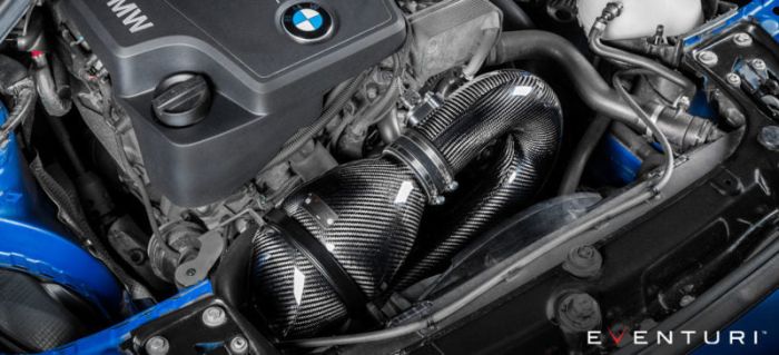 Engine compartment featuring a BMW engine with a carbon fiber intake system by Eventuri, installed and surrounded by various automotive components within a car’s engine bay. Text: “EVENTURI.”