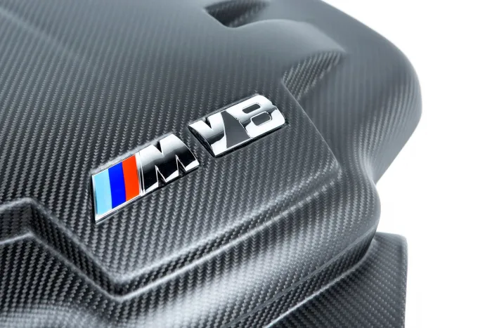 Carbon fiber car engine cover with a glossy "M V8" emblem featuring blue and red stripes, suggesting high-performance, situated in a well-lit environment.
