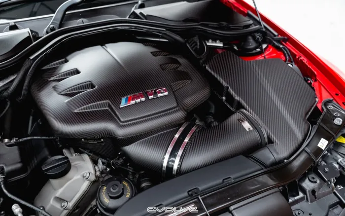 A carbon-fiber engine cover features an "M V8" emblem. The engine is in a clean, organized engine bay within a red sports car. The bottom of the image displays the brand name "EVOLVE."