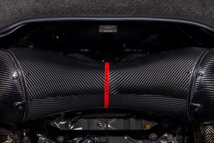 Carbon fiber air intake with a red strap labeled "EVENTURI" is installed in a vehicle engine bay featuring clean, organized components and a gray metallic backdrop. A small label reads "EVENTURI MADE IN ENGLAND."