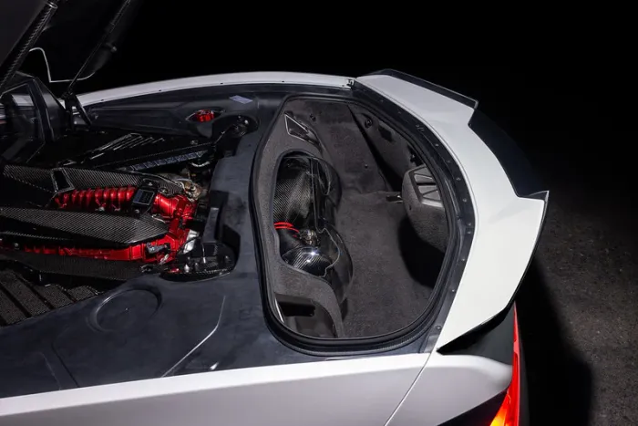 The rear end of a white sports car, with its trunk open, exposing a complex, red, and black carbon-fiber clad engine under the hood, in a dark setting.