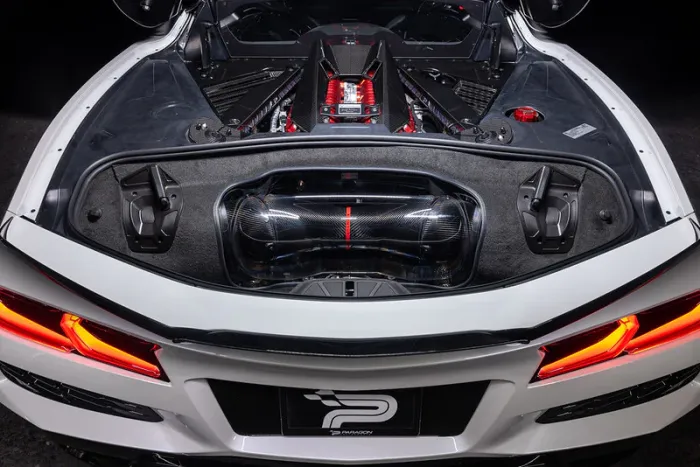 The rear engine compartment of a white sports car displays a high-performance engine with red accents and carbon fiber details under a clear cover, set against a dark background.