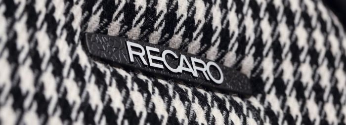The "RECARO" logo is displayed on a black and white houndstooth-patterned fabric, suggesting a close-up view of a seat or upholstery.