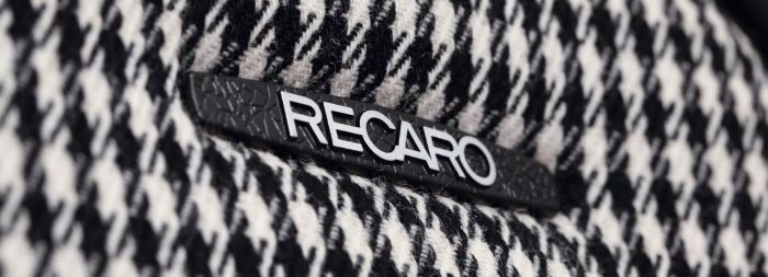 A "RECARO" branded, black nameplate embedded on a black-and-white houndstooth patterned fabric, likely from a piece of automotive or office seating.