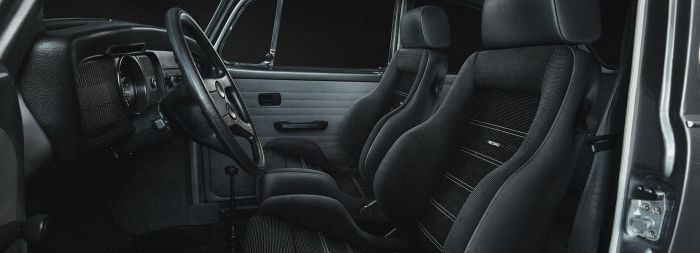 The image features the interior of a car, with black, sporty Recaro seats and a steering wheel. The setting is the dark interior cabin, highlighting vintage automotive design.