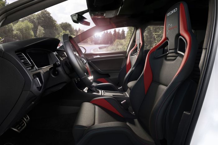 A luxurious car interior featuring black and red Recaro racing seats, a modern dashboard, and a steering wheel with red stitching. The car is on a winding forest road, seen through the windshield. Text on seats reads: "RECARO" and "Nürburgring".