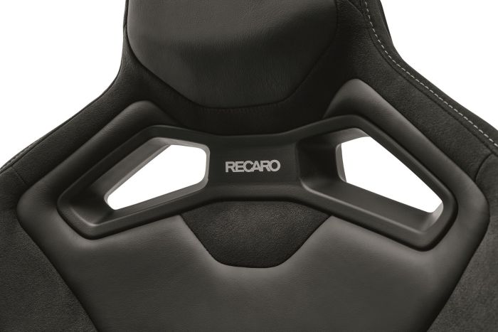 A black, high-backed Recaro car seat with two rectangular cutouts and "RECARO" logo centrally positioned, with detailed stitching visible on the edges.