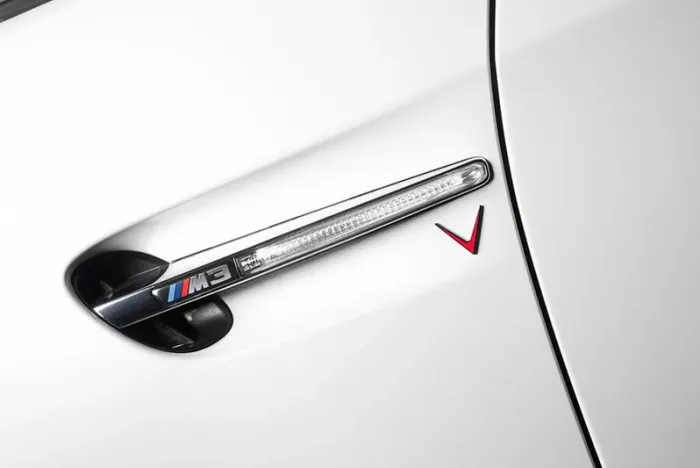 The "M3" emblem and a red V-shaped badge are affixed to the side of a white car, positioned near a sleek side indicator light.