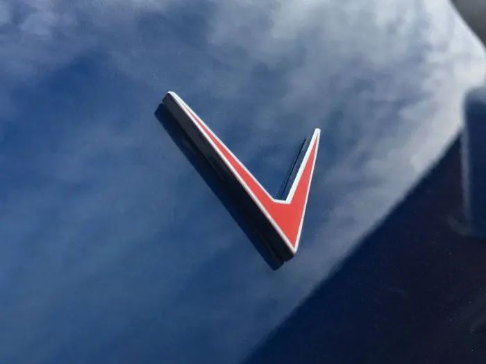 Red, black, and white V-shaped emblem attached to a reflective, shiny blue surface, likely a vehicle exterior. The surrounding area shows blurred reflections and light clouds.
